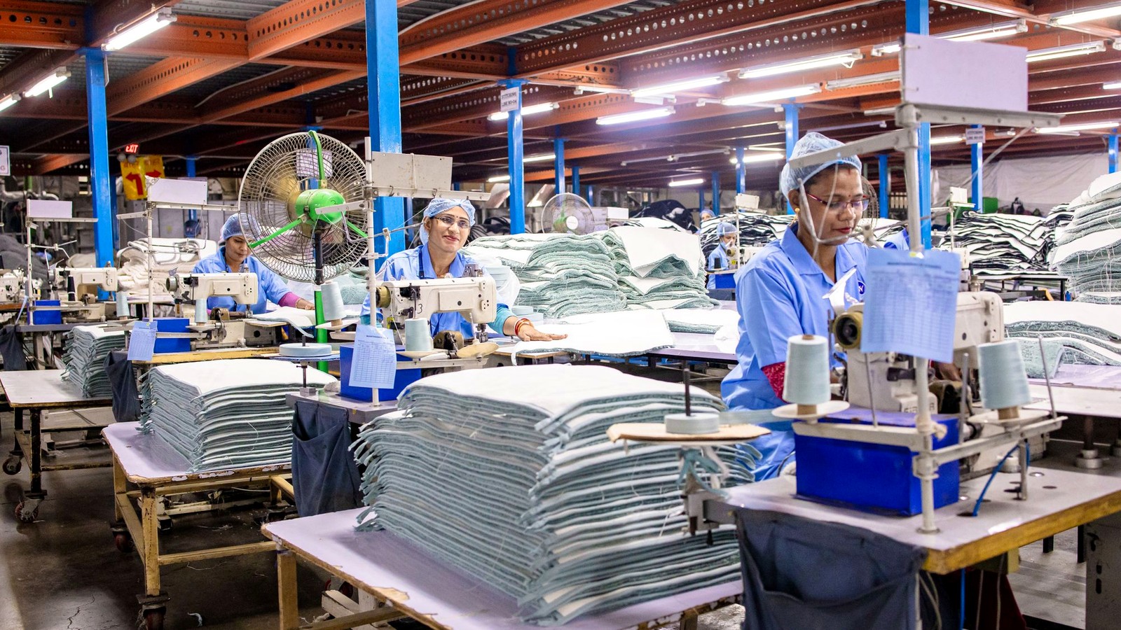 Three workers in red helmets and uniforms inspecting a green fabric bag together on a factory floor, illustrating a quality inspection conversation between factory staff and production workers during a repeat order from China.