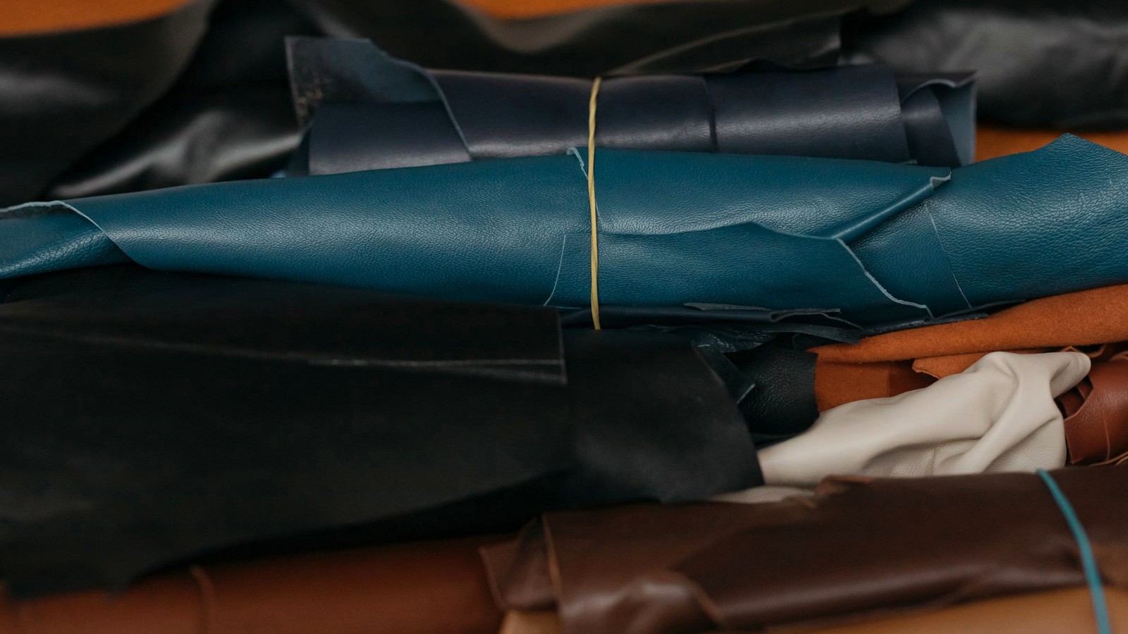 A leather workshop bench with rolled hides in blue, black, brown and cream, illustrating the material choices that can be changed between a first order and a repeat order without altering the appearance of the finished product.