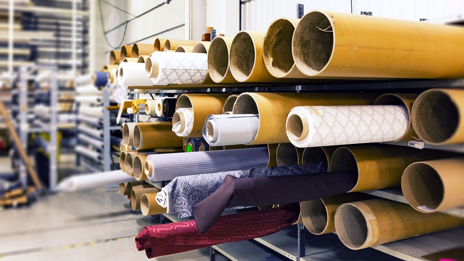 Rows of material rolls stored on racks in a factory warehouse, illustrating how material stock is selected and substituted within a factory.